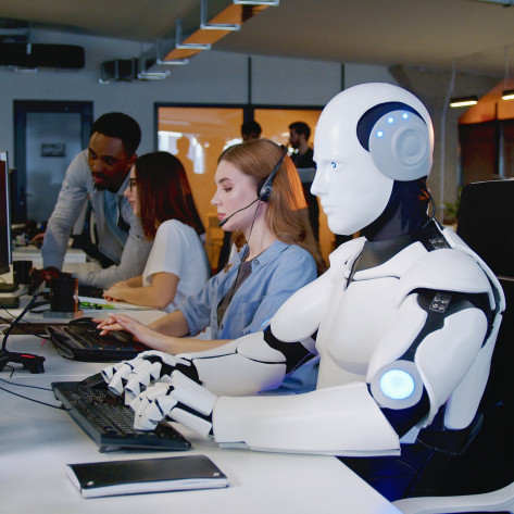 Eine KI sitzt im Büro am PC neben Arbeitskollegen