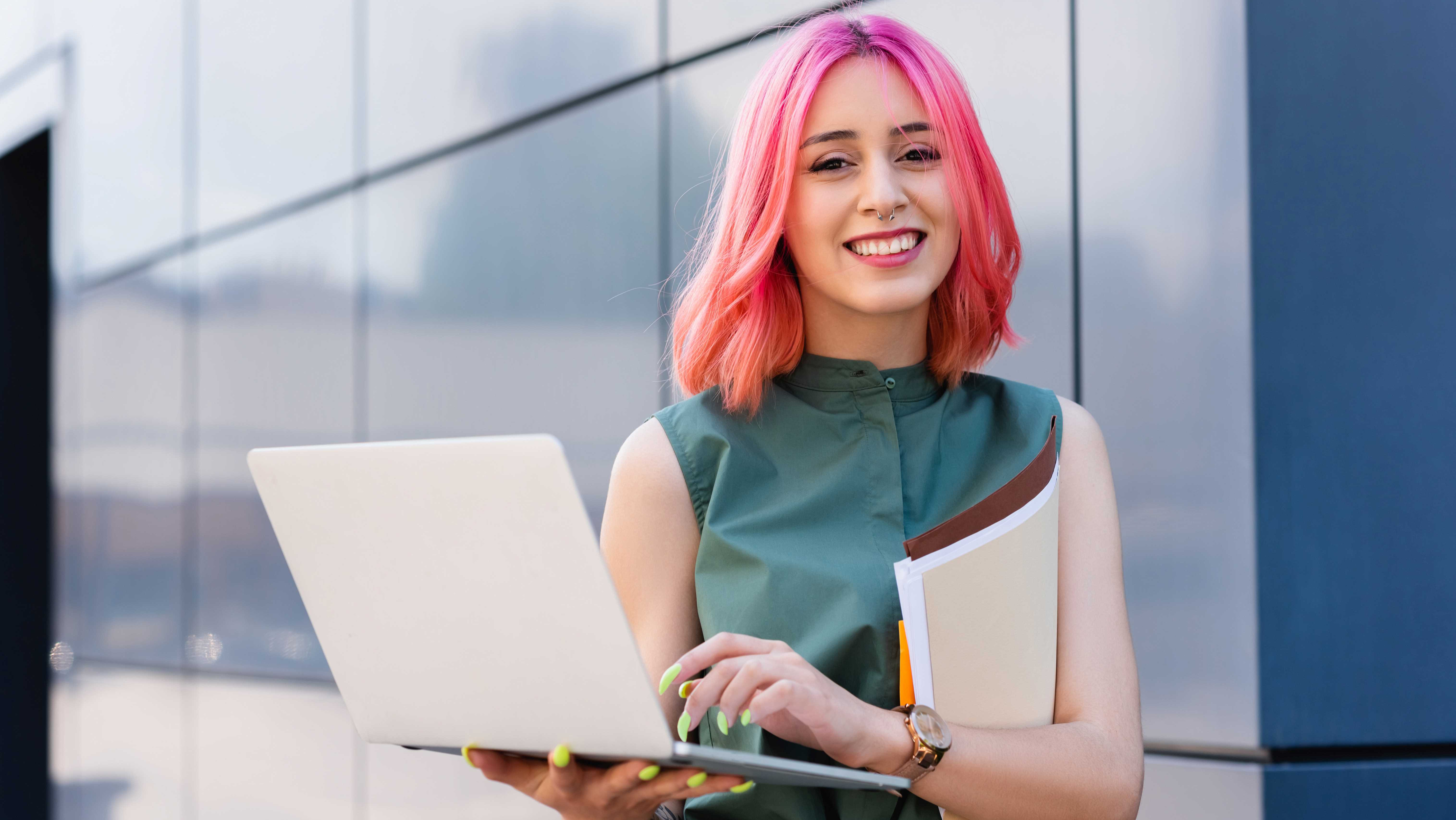 Eine junge Frau mit Pinken Haaren steht mit Ihrem Laptop da